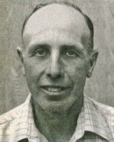 A black and white portrait of a man with short hair wearing a checkered shirt, smiling at the camera.