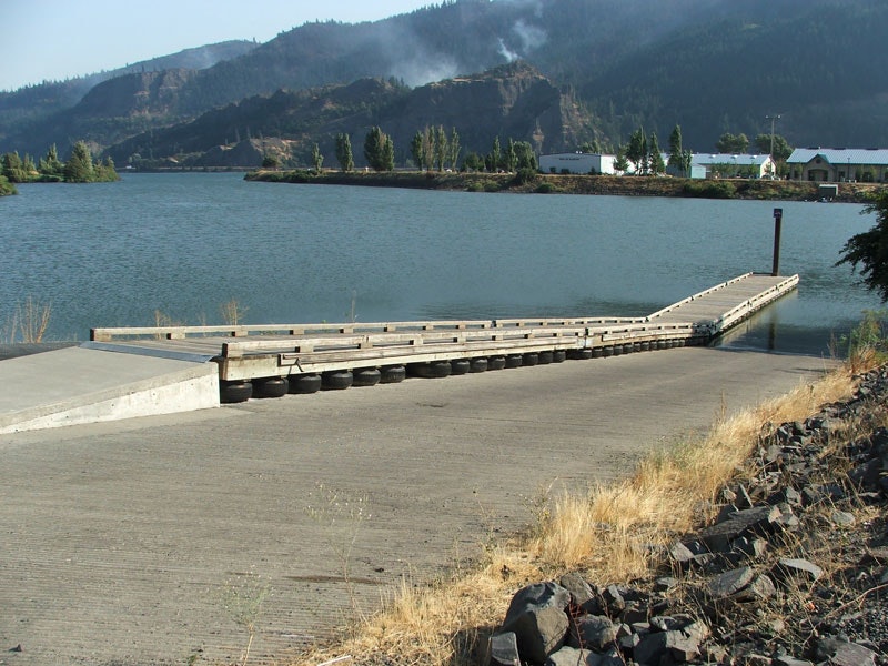 Boat ramp leading to a calm river, mountains and smoke in the background, rocky path on the right.