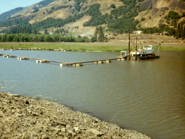 A barge connected by a long floating structure on a river, with a hilly, wooded background.