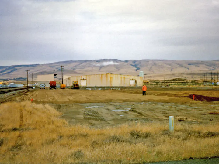 Wide open field with a few vehicles and buildings in the background; mountains under cloudy sky.