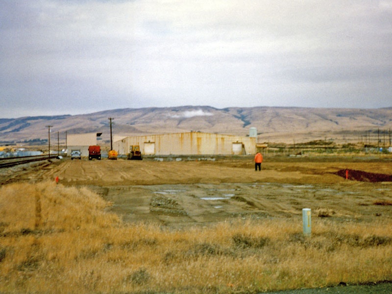 Wide open field with a few vehicles and buildings in the background; mountains under cloudy sky.