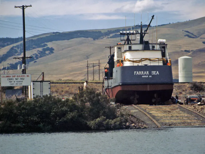 A ship labeled "Farrar Sea" is on land near a body of water, with hills in the background and a sign for Columbia Shipbuilders.