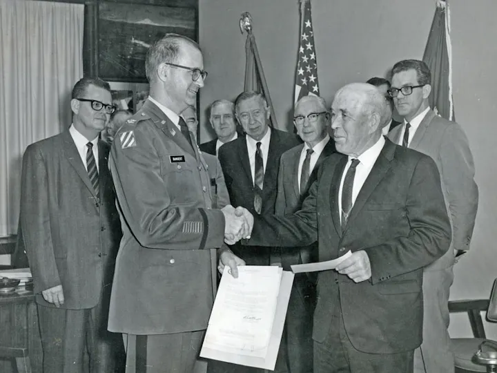 A military officer shakes hands with a man in a suit, surrounded by onlookers and flags.