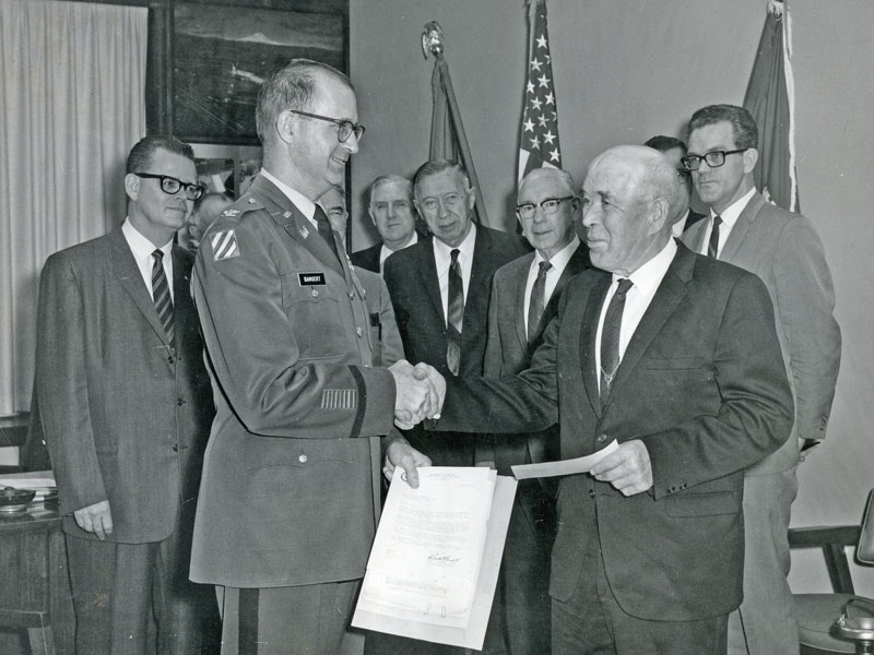 A military officer shakes hands with a man in a suit, surrounded by onlookers and flags.