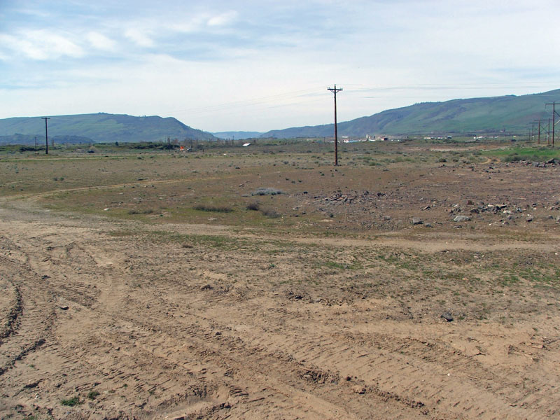 A dry, empty field with utility poles and distant hills under a partly cloudy sky.