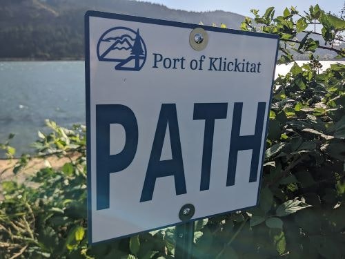 Sign reading "Port of Klickitat PATH" by a natural area with water and greenery.