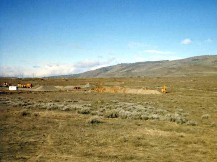 A barren landscape with construction equipment, dirt piles, and distant hills under a blue sky.