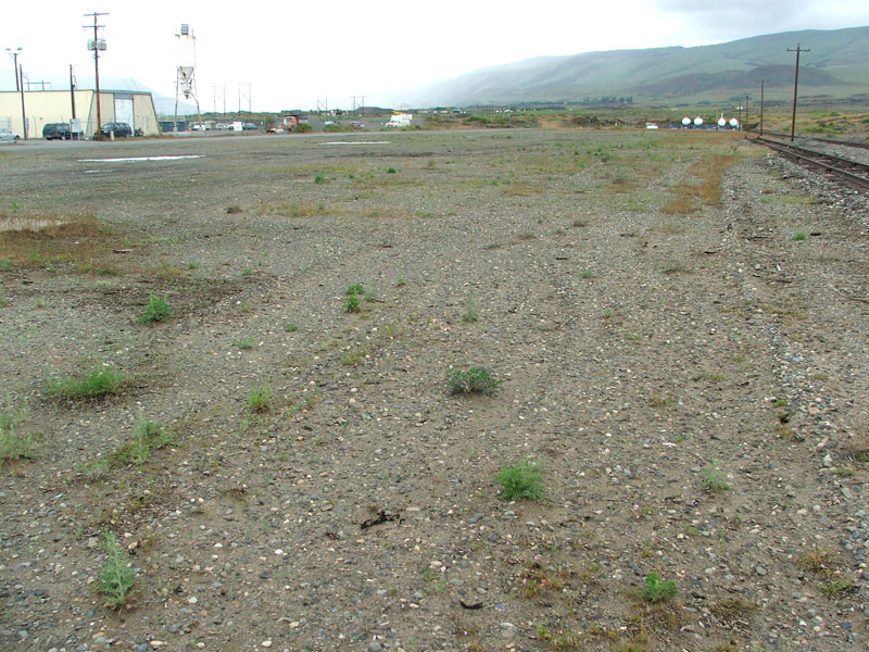 Gravel lot with sparse vegetation, adjacent to railway tracks with industrial buildings and hills in the background.