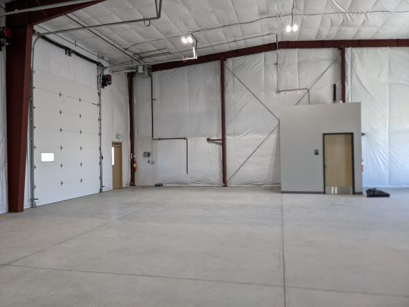 Empty warehouse interior with white paneled walls, large garage door, small room with door, and concrete floor.