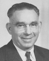 A black-and-white portrait of a man in a suit and tie, wearing glasses, smiling.