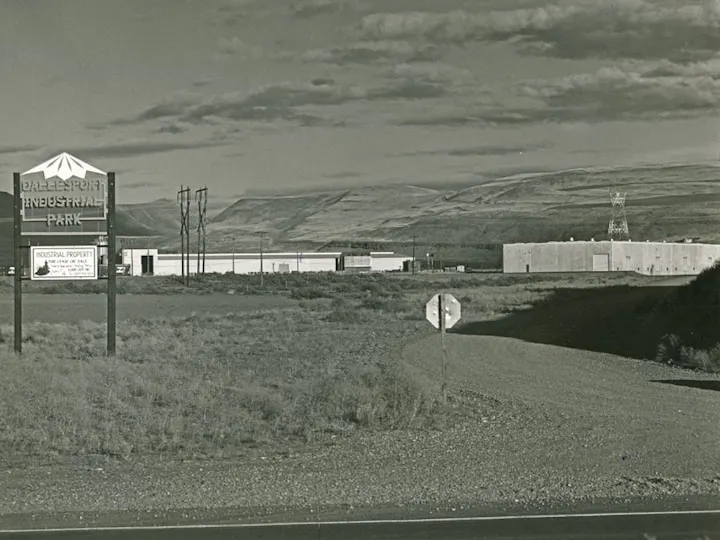 Industrial park sign and warehouses in a desolate landscape with hills in the background under a cloudy sky.