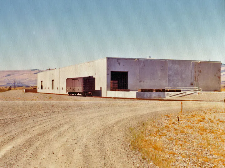 A large, rectangular industrial building with a boxcar on a gravel path in a desert landscape under a clear blue sky.
