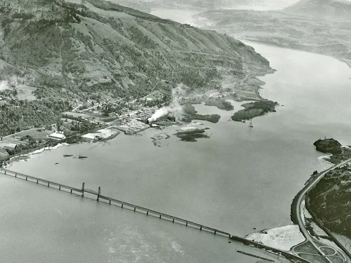 Aerial view of a river with a bridge, hills, and an industrial area emitting smoke.