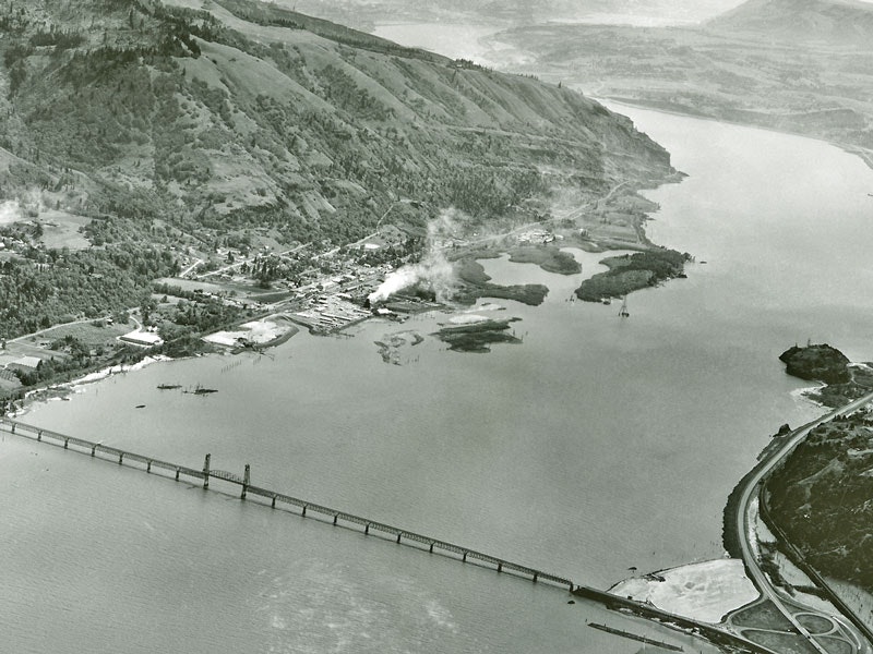 Aerial view of a river with a bridge, hills, and an industrial area emitting smoke.