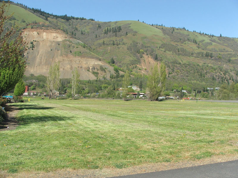 A grassy field with trees, a distant hilly landscape, and a few buildings under a clear blue sky.