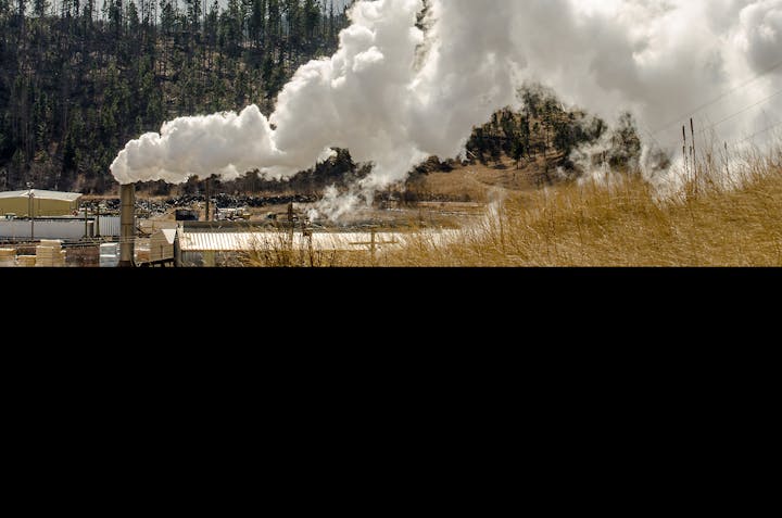 Industrial facility emitting large plumes of steam or smoke, with forested hillside in the background.