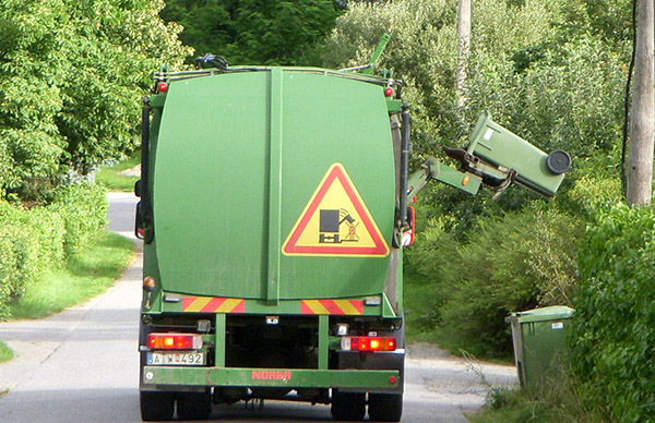 A green garbage truck with an automated arm lifting a trash bin on a tree-lined road.