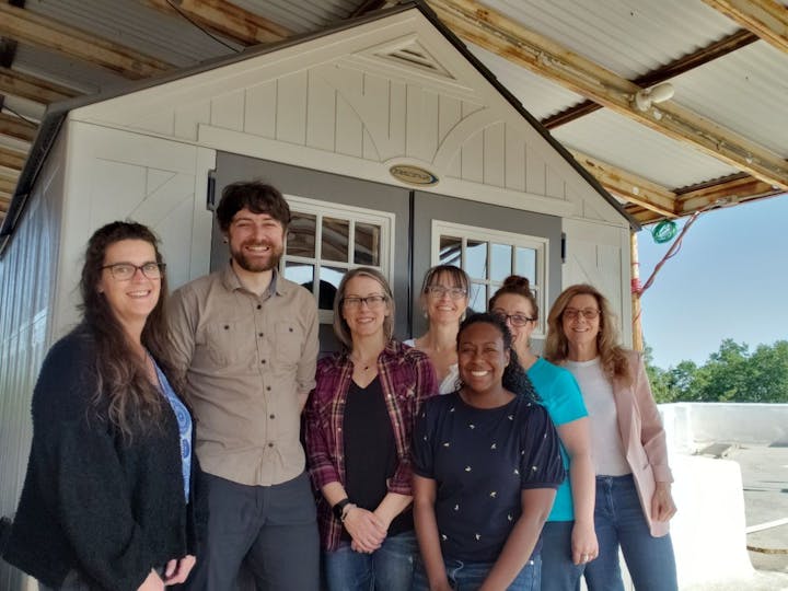 A group of seven people smiles together in front of a small shed or structure under a roof.