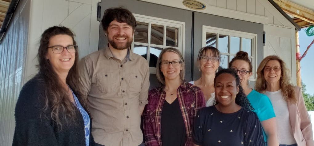 A group of seven people smiles together in front of a small shed or structure under a roof.