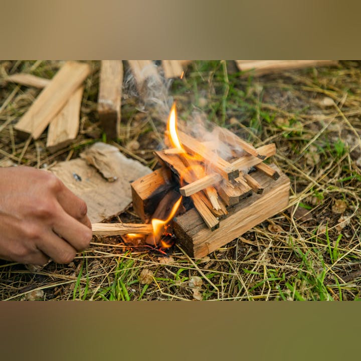 A person is starting a fire using kindling and a stick, with flames and smoke rising amidst grass and wood.