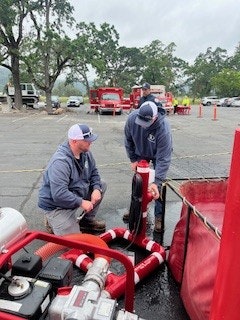 Three individuals are working with firefighting equipment in an outdoor setting, likely preparing for a training or response scenario.