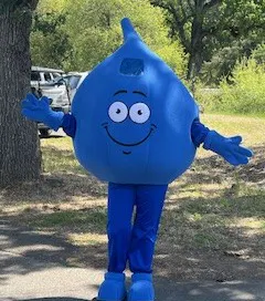 A friendly blue water droplet mascot with a big smile, standing outdoors and inviting interaction.