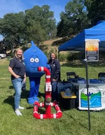A blue water droplet mascot poses with two women at an outdoor event, alongside a booth and a water pipe display.