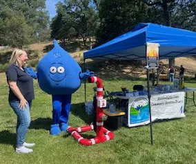 A cheerful blue water droplet mascot stands next to a woman at a booth with a tent promoting water conservation.