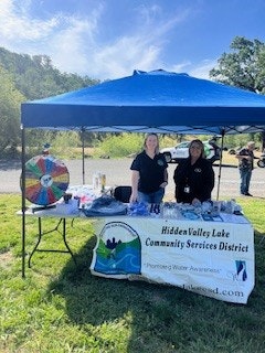 Two women stand behind a booth with a blue tent, promoting Hidden Valley Lake Community Services, with a spin wheel and various materials.