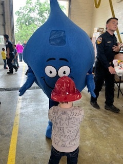 A child wearing a gray shirt stands next to a blue cartoon water droplet mascot, both smiling, with people in the background.