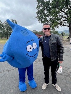 A person poses with a large blue cartoon water droplet character at an outdoor event.