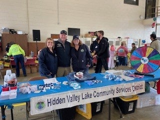 The image shows three people at a table promoting Hidden Valley Lake Community Services, with a colorful prize wheel and various items.