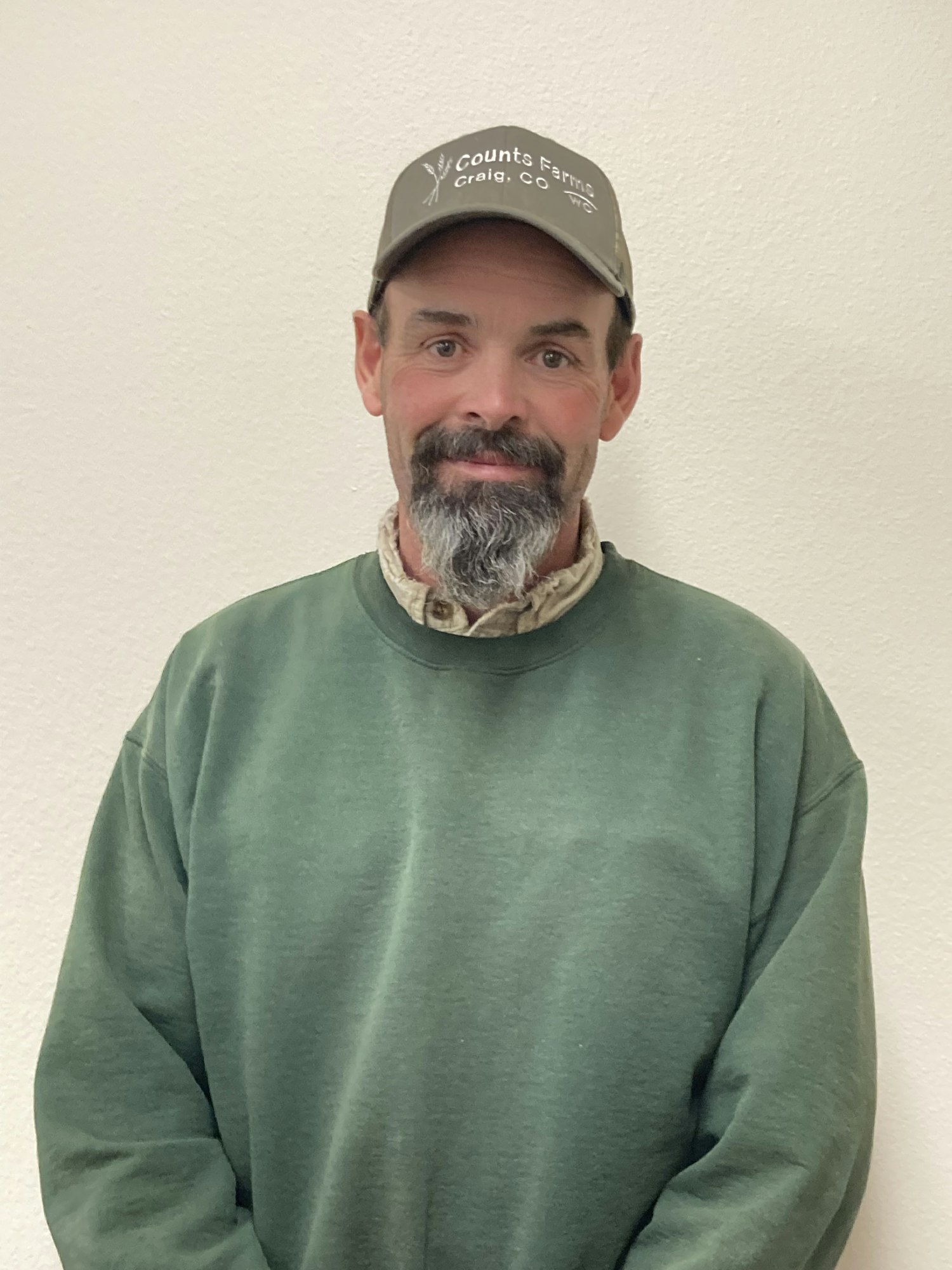 Man in a green sweatshirt and cap, with beard, smiling at the camera.