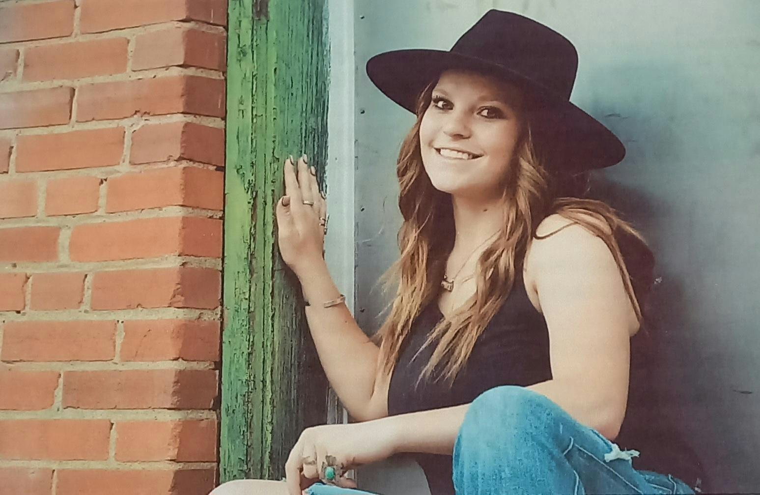 A woman in a black hat smiles while seated against a wall, with a hand on a green wooden door next to a brick wall.
