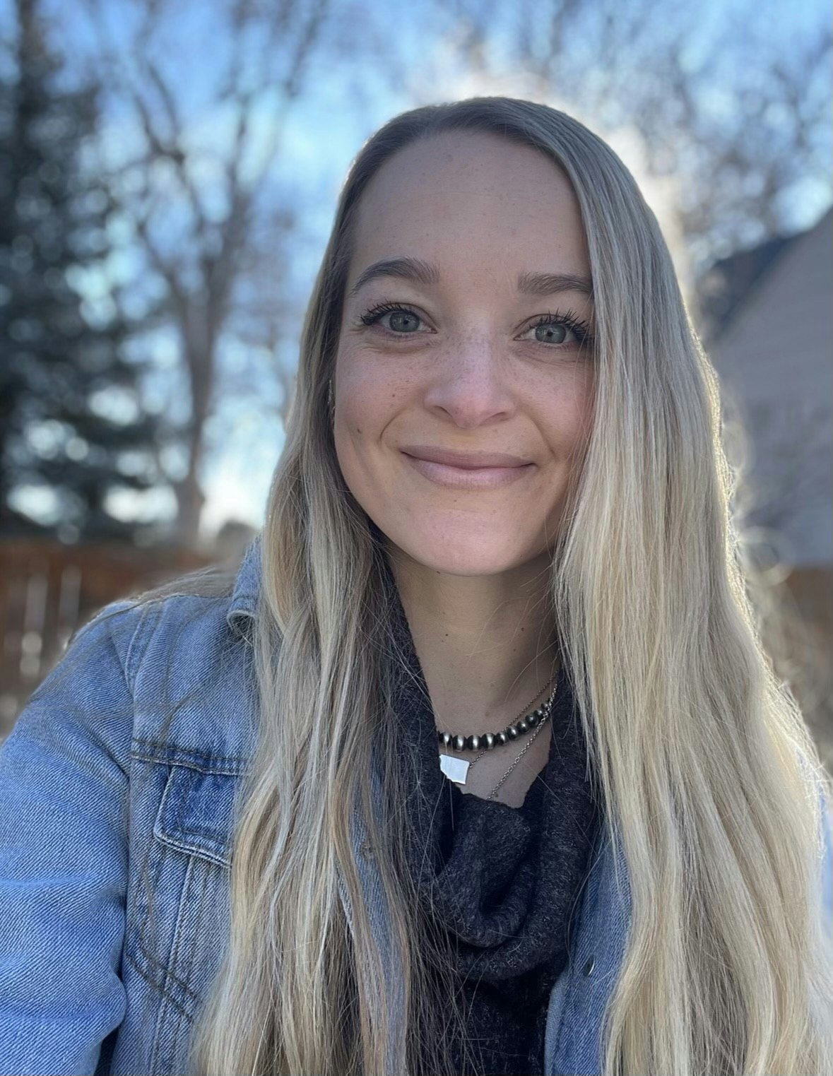 A person with long hair, wearing a denim jacket, smiles outdoors with trees and a house in the background.
