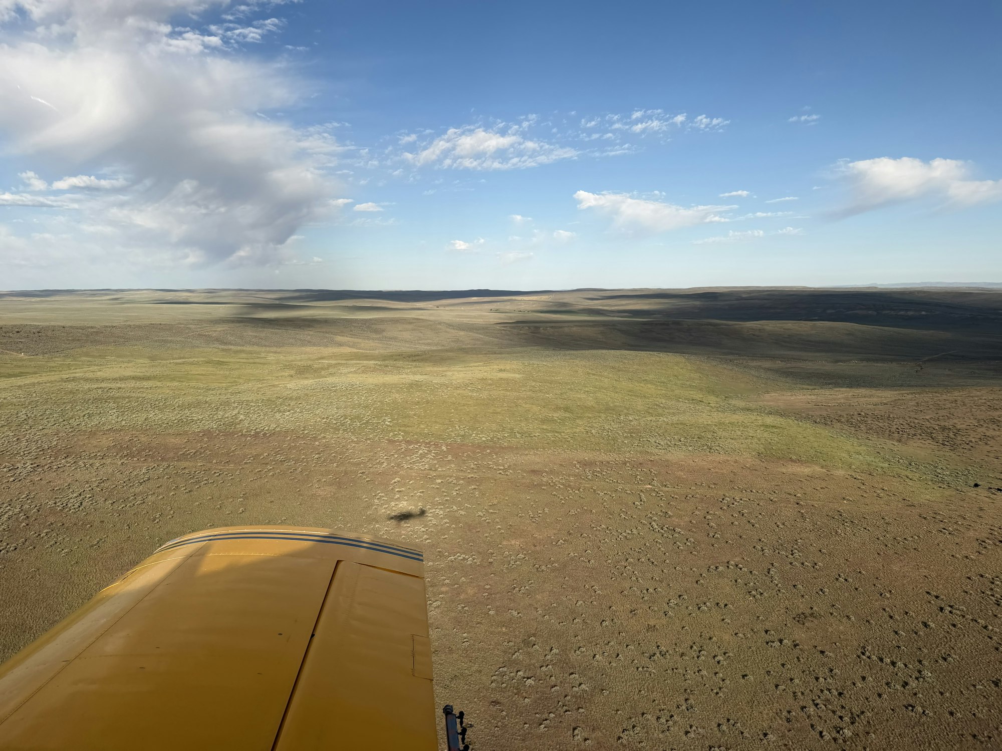 Aerial view of a vast, open landscape with grasslands under a blue sky with scattered clouds, featuring the wing of a yellow plane.