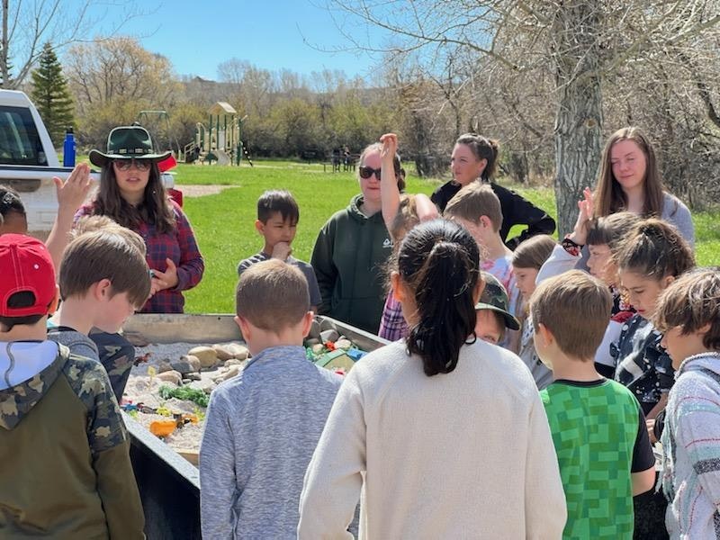 A group of children and adults gathered outdoors around a table with various items, possibly for an educational activity.