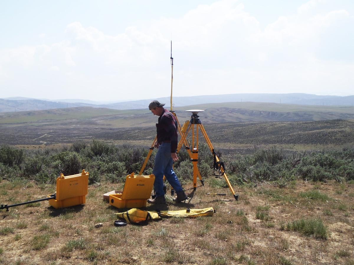 A person using surveying equipment in an open, hilly landscape.