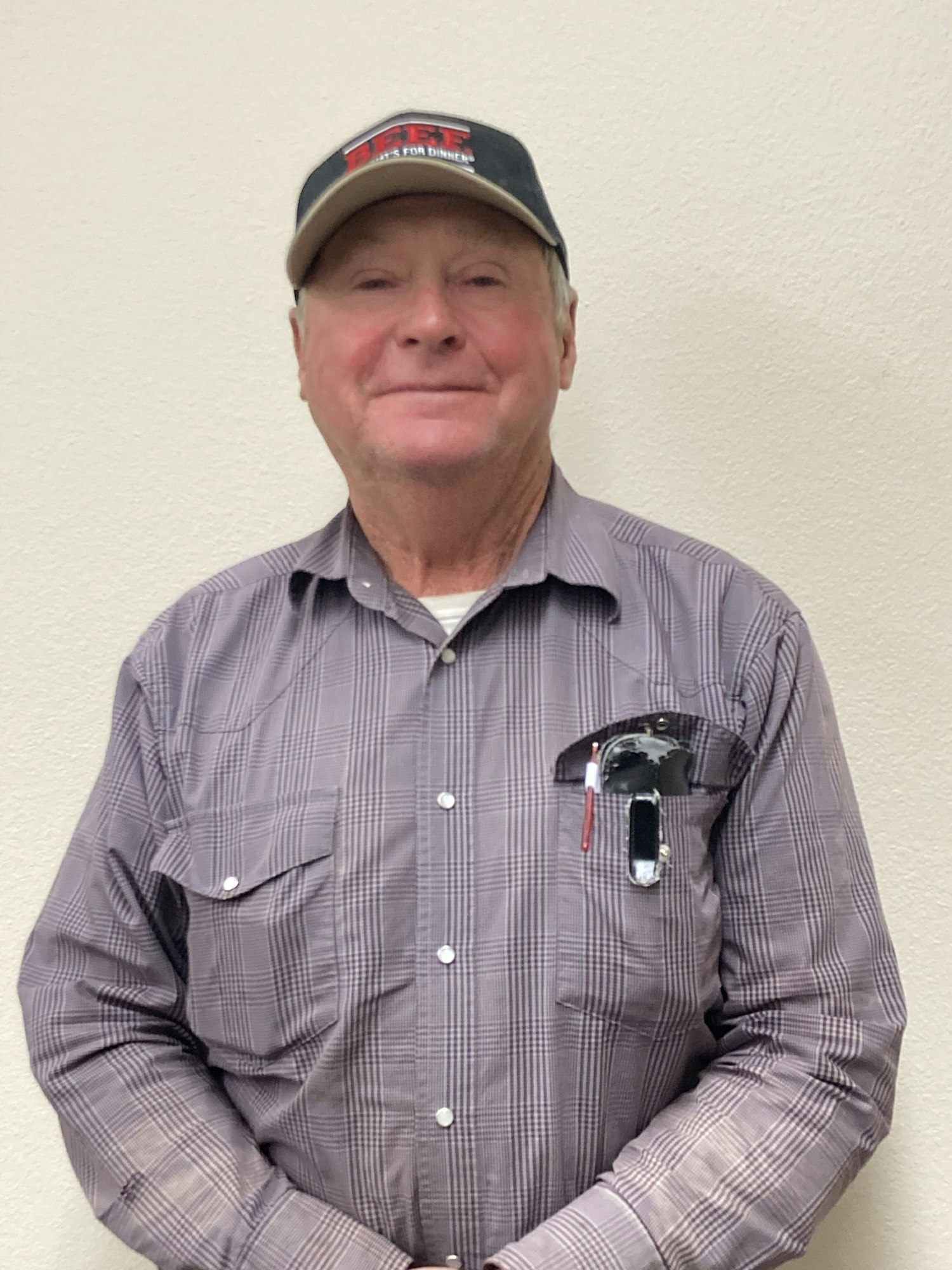 Man in a plaid shirt and baseball cap, sunglasses in pocket, standing against a light background.
