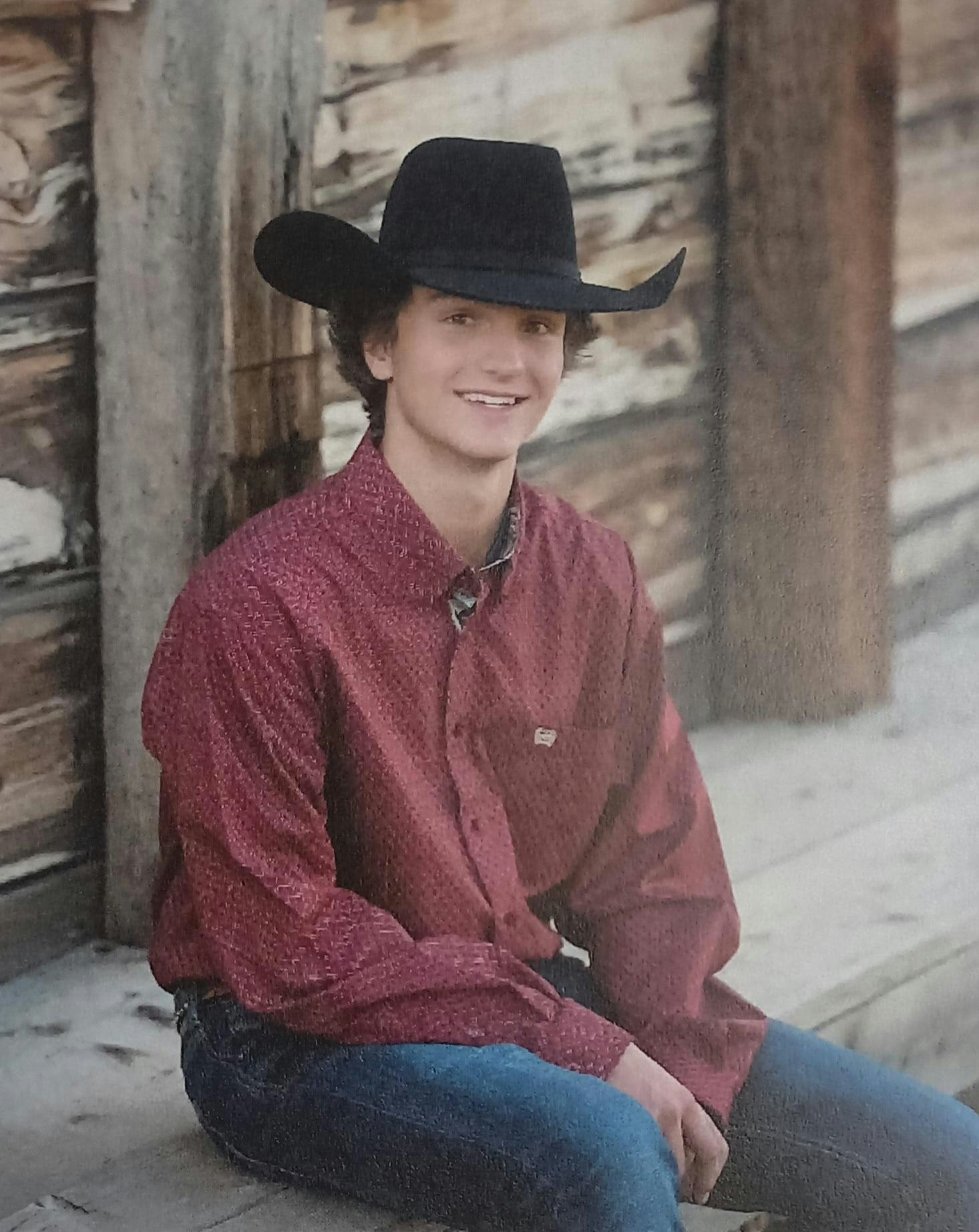 A person in a cowboy hat, red shirt, and jeans, sitting and smiling next to a wooden structure.