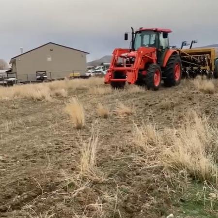 A red tractor pulling a large attachment across a dry field near a building.