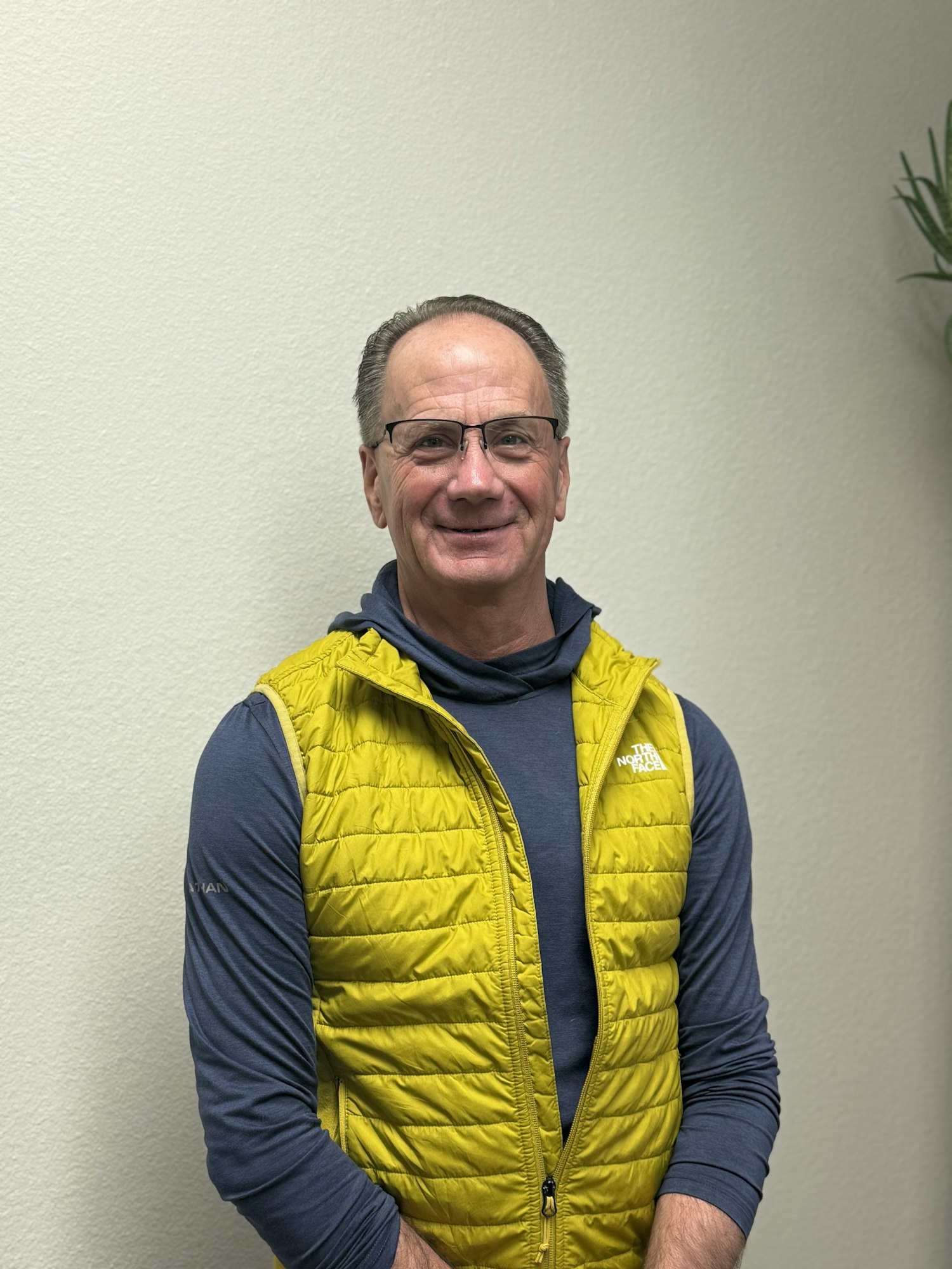 A person wearing glasses, a bright yellow vest, and a blue shirt, smiling against a plain wall.