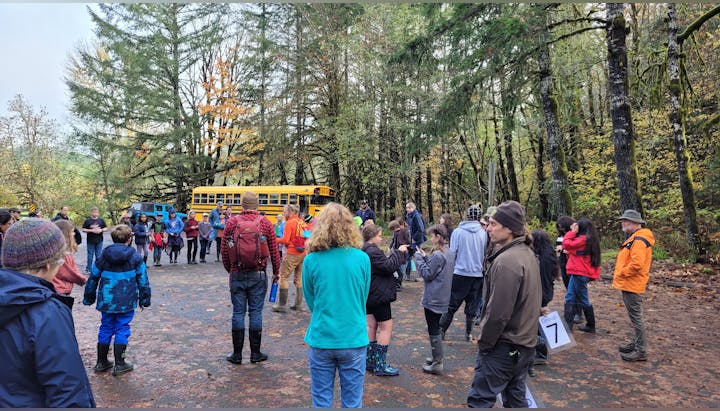 A group of people, possibly on a field trip, gathered in a forested area with a yellow school bus nearby.