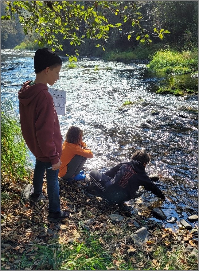 Three people by a river, one standing, two sitting, surrounded by nature and sunlight.