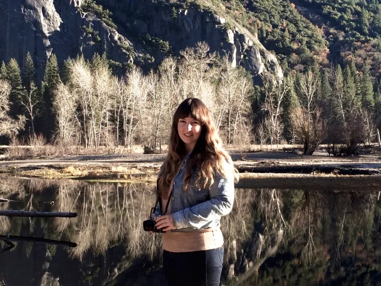 A person holding a camera stands by a reflective body of water, with trees and rocky cliffs in the background.