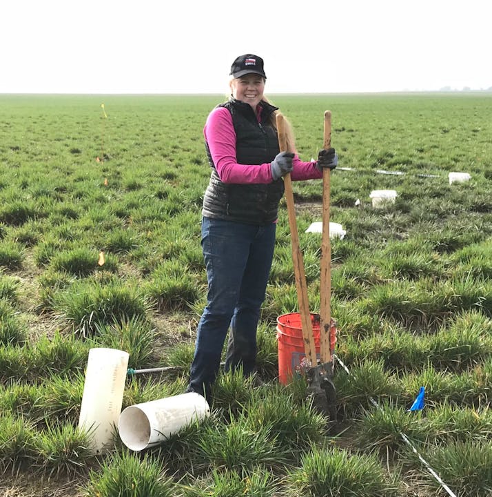 A smiling person in a field with a shovel, a bucket, and white tubes on the ground.