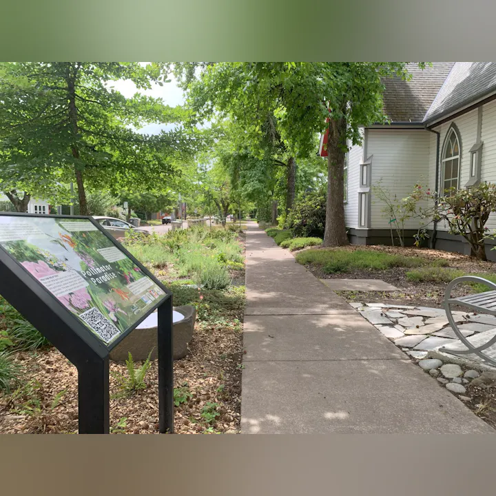 The image shows a sidewalk beside a garden with a sign about a "Pollinator Paradise," surrounded by trees and a building.