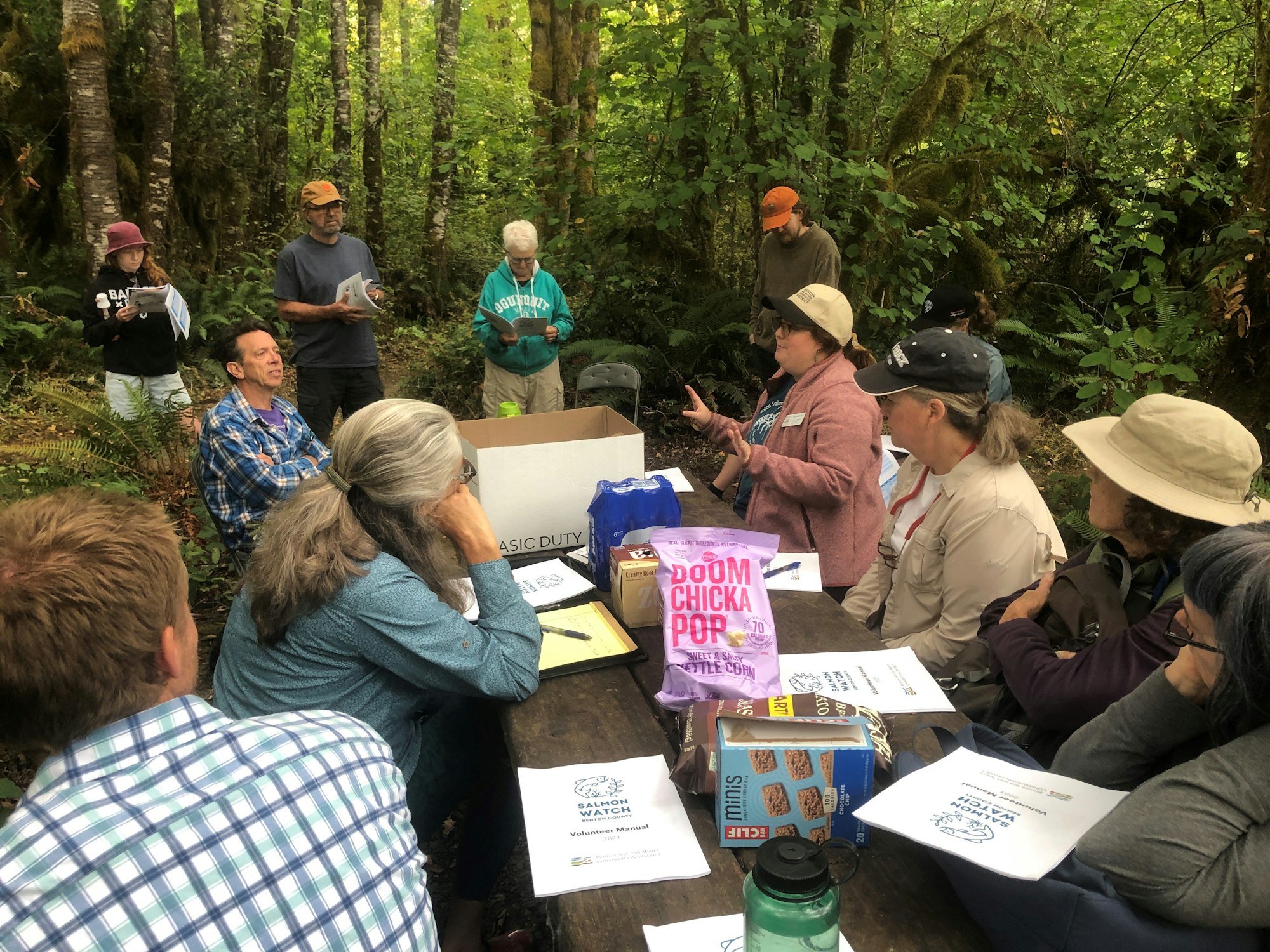A group of people having a meeting outdoors in a forest, with snacks and papers on a table.