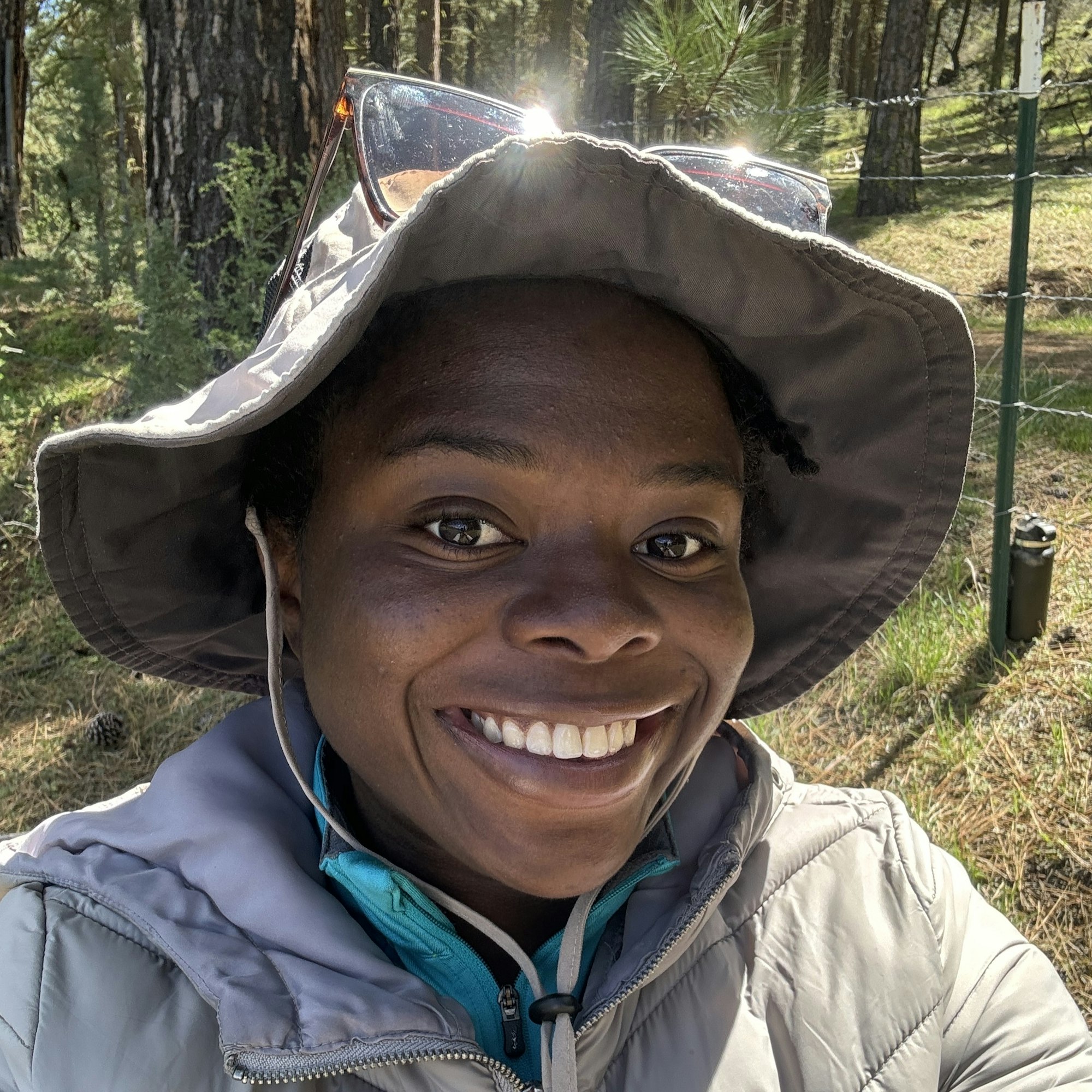 A smiling person with a hat and sunglasses outdoors.