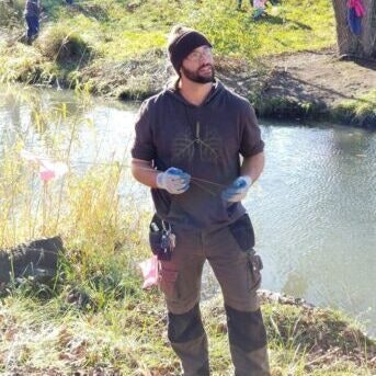A person outdoors near a river, wearing gloves and a hat, holding a stick or rod, dressed in work clothes.